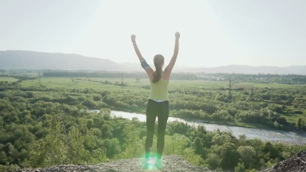 Молодая спортивная девушка, стоящая на вершине высокой скалы над рекой на прекрасном золотом закате, поднимая руки вверх, весело подпрыгивает. Молодая спортсменка стоит на вершине — стоковое видео