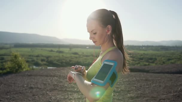 Atleta di fitness ragazza che corre veloce con velocità indossando bracciale del telefono mostrando con smartphone touch screen sulla montagna al tramonto. Runner che ascolta musica. Allenamento cardio — Video Stock