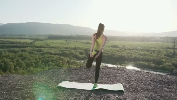 Het meisje sport voert yoga oefeningen in de outdoor. Achterkant weergave slim blonde meisje staat in yoga vormen Vrikshasana op groene mat in de buurt van knipperen onder zonlicht meer — Stockvideo