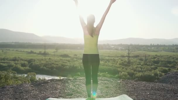 Vista trasera. Joven deportista practicando yoga en la cima de la montaña al atardecer. Mujer delgada haciendo gimnasia sobre el sol de la mañana. Chica con una figura delgada practica yoga cerca del río en la cima de la montaña en — Vídeo de stock