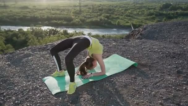 Yoga of turnster jonge vrouw in Romper beoefenen van yoga bij fantastische zonsopgang of zonsondergang. Fitness, sport, yoga en gezonde levensstijl concept. Meisje doet een brug boven op de berg boven de rivier — Stockvideo