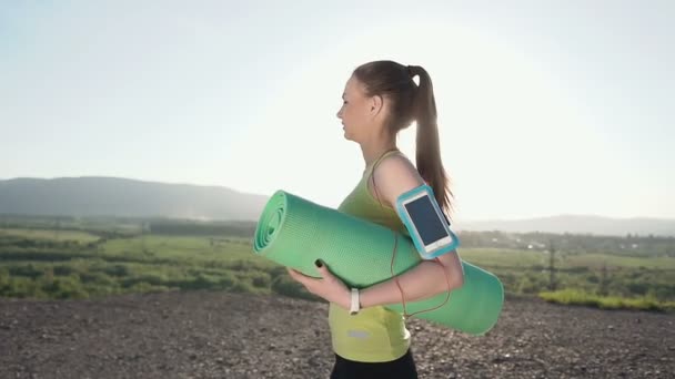 Молодая спортивная женщина ходит на вечернюю тренировку на закате. Девушка держит зеленый коврик для йоги. Спортсмен проходит по вершине горы с ковриком для занятий — стоковое видео