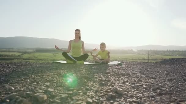 Pequeño hijo y madre haciendo yoga y meditación al aire libre en la cima de la montaña en el sol del este por la mañana. Entrenamiento deportivo en la naturaleza. Hermana y su hermano menor realizan yoga — Vídeos de Stock