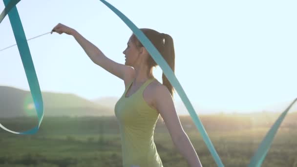 Fecha. Ginasta feminina atraente dançando com fita azul ao ar livre ao nascer do sol ou ao pôr do sol. Ginasta magro em corpo verde realiza o exercício de ginástica com uma fita azul na natureza — Vídeo de Stock