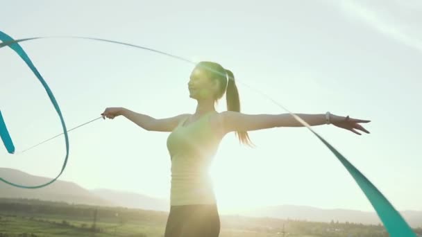 Rhythmische Sportgymnastik: Mädchen im Sportbody turnen im Freien bei Sonnenuntergang oder Sonnenaufgang mit einem blauen Band. Turnerische Übungen — Stockvideo