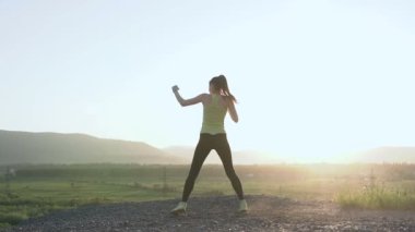 Egzersiz dağın zirvesinde gündoğumu veya günbatımı yapıyor boxings kız o doğada yüksek tekme egzersiz. Kadın kendine güvenen boksör ile mavi gökyüzü karşı bir tutum sergilemek savaşmak. Muzaffer spor kadında