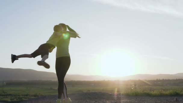 Ein glücklicher Sohn und eine glückliche Mutter. Eine fröhliche Sportlerin spielt mit dem Kind bei Sonnenuntergang nach dem Lauftraining in der Natur. Die junge Mutter umkreist das Kind, indem sie seine Hände nimmt. Gegenlicht. schön — Stockvideo