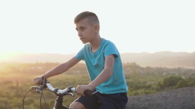 Yakışıklı bir çocuk 8-9 eski mavi bir tişört gündoğumu ve günbatımı orman arka planı dağlık bir alanda bir bisiklet sürmek. Yaz başlangıcında, sportif siyah Bisiklete binmek bir çocuk