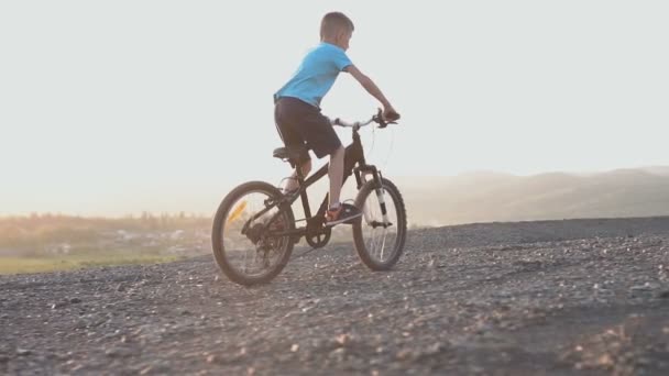 Vista trasera de un niño de 8-9 años que monta una bicicleta deportiva negra en una zona montañosa en el verano al atardecer. El chico está vestido con una camiseta azul y pantalones cortos negros. Paseo en bicicleta al amanecer — Vídeos de Stock