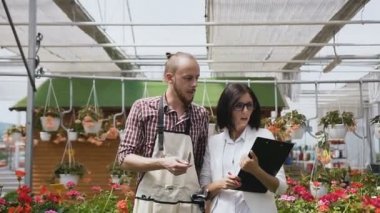 Genç insanlar işçiler tarım sektörünün git sera ve süs bitkileri inceleyin. Bahçıvanlar, adam ve kız çiçekler sera içinde saymak