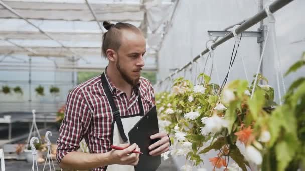 Ein junger Gärtner mit Bart und Schürze führt Buch über Zierpflanzen. Tageslicht natürliches Licht. — Stockvideo