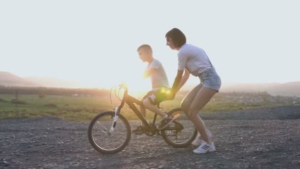 Una joven madre enseña a su hijo a conducir en bicicleta en verano al atardecer o al amanecer. Una mujer sostiene una bicicleta en la que su hijo aprende a montar felices vacaciones familiares, montando en bicicleta — Vídeos de Stock