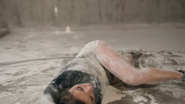 Dançarina menina executa uma dança moderna deitada no chão em uma nuvem de pó ou farinha ou poeira. Jovem mulher em um terno de corpo preto dança contemporânea no chão no estúdio — Vídeo de Stock