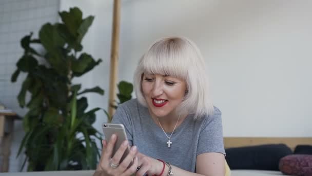 Flicka leenden när du tittar på ett foto på sin telefon. En kvinna i sängen på bakgrunden av en vit vägg och en stor skål är roligt tittar på sin smartphone. — Stockvideo
