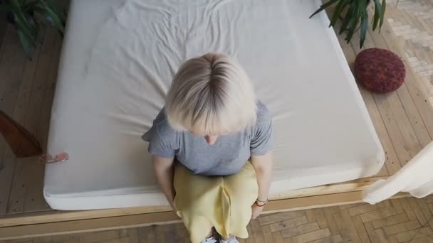 Vue de dessus, beau plaisir blonde se trouve dans le grand lit et se brise les bras. Fille souriante dans un lit blanc sur un fond de grandes plantes vertes. Le concept de la chambre du matin de bonheur et de joie . — Video