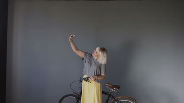 Portrét krásné dívky sedí na kole a fotografování sama na telefonu. Blondýna na kole díky selfie v jasné místnosti. — Stock video