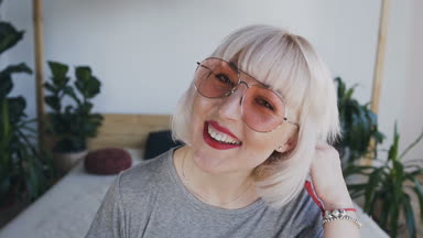 Kamera seyir bir yatak üzerinde oturan genç sarışın bir kadın. bir kız gözlük ve beyaz bir duvar arka plan üzerinde bir yatak odasında bir dövme.