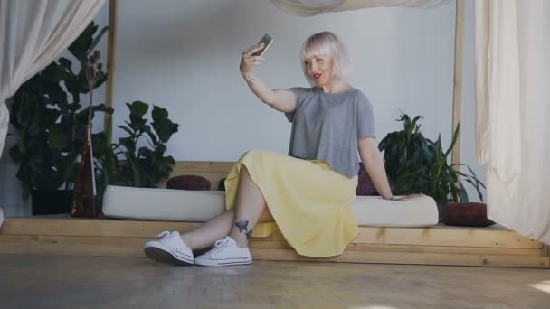 Algemeen plan. Een meisje met witte haren zit op de rand van een mooi bed en pakt op de telefoon. Celphy vrouw in een lichte slaapkamer. — Stockvideo