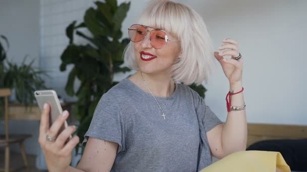 Close-up Una mujer con gafas naranjas y tatuajes en la mano mueve su pelo blanco y recoge su teléfono. Una chica sonriente y feliz en una gran cama blanca . — Vídeo de stock