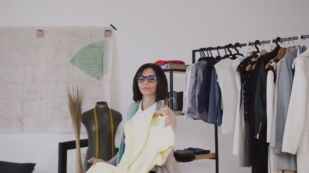 Une brune heureuse fait une robe jaune ajustée, qui est sur un cintre dans le studio de créateurs de mode. Vêtements, garde-robe, mode, style et concept des peuples — Video