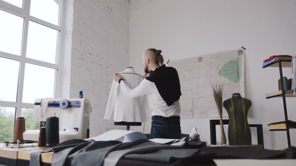 Молодой креативный портной измеряет белый пиджак на манекете в своей мастерской для шитья одежды. Бородатый модельер измеряет новую модель одежды, которую он производит, танцуя — стоковое видео