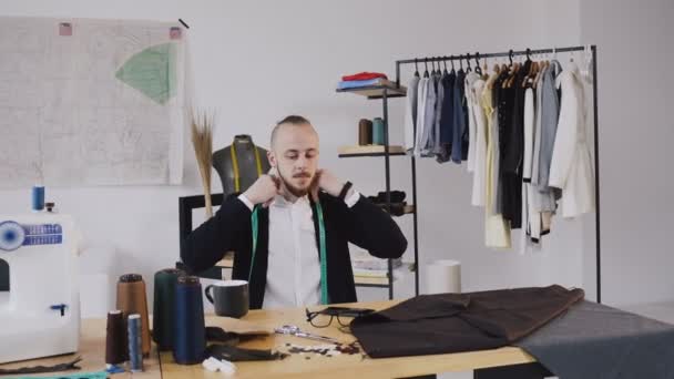 El joven sastre caucásico está tomando café caliente antes de empezar a trabajar por la mañana. En un estudio de diseño de moda y sastrería bebe café mientras piensa en el desarrollo de una nueva colección — Vídeos de Stock