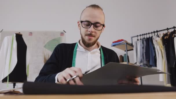 Primer plano de un joven diseñador barbudo o sastre con gafas que se sienta en una mesa de trabajo en el estudio y esculpe con tijeras unos bocetos para coser una nueva colección de ropa. Un barbudo con estilo — Vídeo de stock