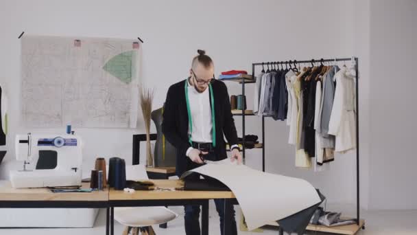 Junger männlicher Designer mit Tape-Line um den Hals steht im Schneideratelier und schneidet mit der Schere ein Muster für eine neue Kollektion. männlicher bärtiger Modedesigner und Schneider im Atelier schneidet ein — Stockvideo