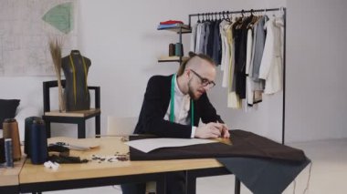 Genç Moda Tasarımcısı beraberlik skeçler gelecek giyim için kumaş kesip cetvel kullanarak sakallı. Kumaş, desen ve dikiş araçları yalan önünde dikiş masasında bir terzi ve genç tasarımcı. Küçük