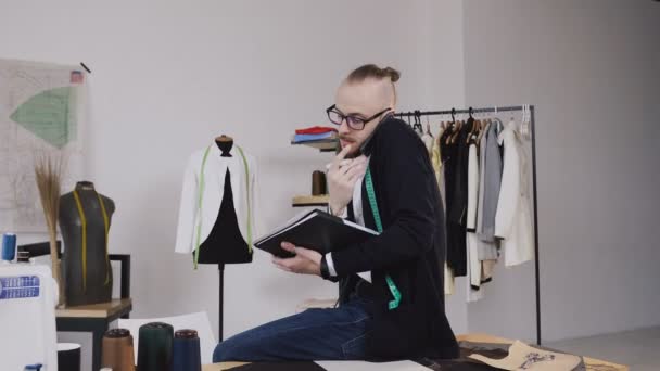 Young fashion designer or tailor sitting on table in atelier and talking phone and makes notes in his notebook. Young bearded man in glasses dressed in white shirt and black blouse with measure tape — Stock Video