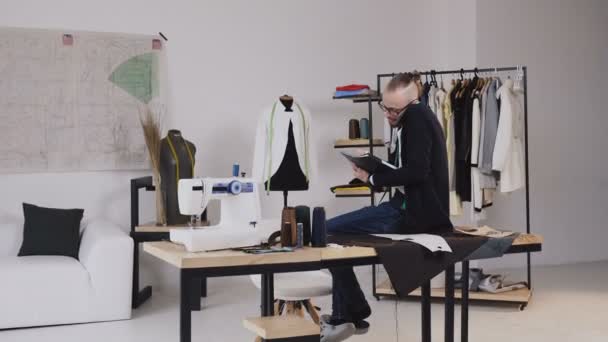 Guapo diseñador de moda sentado en la mesa en el taller y hablando de teléfono, toma notas en su cuaderno. Joven sastre barbudo en gafas vestidas con camisa blanca y blusa negra con cinta métrica — Vídeos de Stock