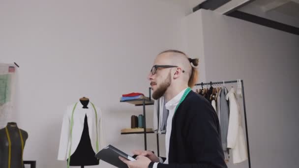 Ein gutaussehender Modedesigner sitzt auf dem Tisch im Atelier und zeichnet Skizzen für die neue Kleiderkollektion. ein junger bärtiger Schneider mit Brille, bekleidet mit einem weißen Hemd und einem schwarzen — Stockvideo