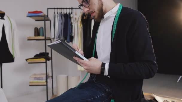 El diseñador se sienta sobre la mesa y dibuja bocetos en el Studio. Diseñador dibujando bocetos para la ropa. Guapo barbudo diseñador trabajando en atelier, bosquejando un proyecto de moda — Vídeo de stock