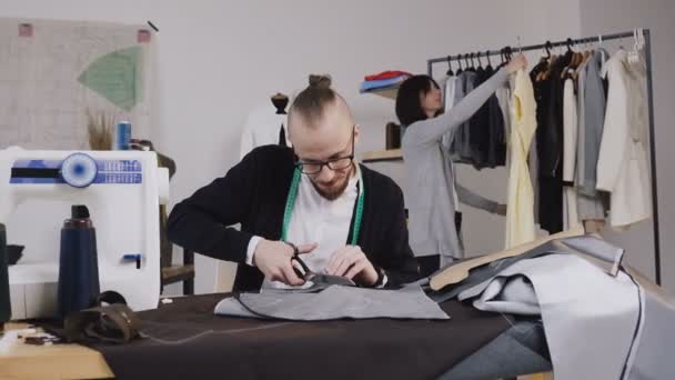 Schneider oder Modedesigner sitzt am Arbeitsplatz im Atelier und schneidet Stoff mit einer großen Schere oder Schere, während er den Kreidemarkierungen des Musters anhand von Skizzen folgt. — Stockvideo