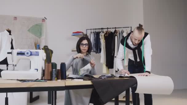 Casal de designers de moda ou alfaiate e costureiro trabalhando com esboços de tecido e roupas no estúdio cheio de ferramentas e equipamentos de alfaiataria — Vídeo de Stock