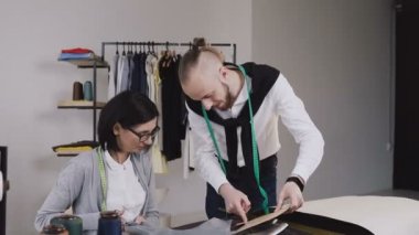 Çok etnik gruptan oluşan moda tasarımcıları desen ve kumaşlar ile farklı terzilik araçları ve elbise ile güzel ofiste çalışan iki kişi. Takım iki terzi gri kumaş ve skeçler, çalışma