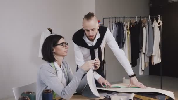 Deux couturiers caucasiens sur mesure travaillant avec des motifs en tissu assis dans le beau bureau avec différents outils et vêtements de couture. Tailleur et couturière sur le lieu de travail au studio — Video