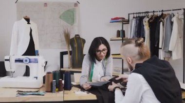 Genç kadın ve erkek moda tasarımcıları veya tablo farklı terzilik araçlarıyla güzel ofiste oturan gri kumaş ile çalışma terziler. İki kişi moda tasarımcıları, kumaş seçimi