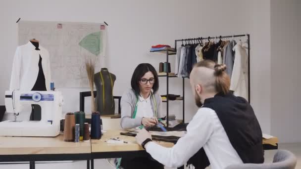 Duas pessoas designers de moda escolhendo tecido no escritório com diferentes ferramentas de costura e roupas. Jovem mulher e homem estilistas ou alfaiates trabalhando com tecidos sentados na bela — Vídeo de Stock