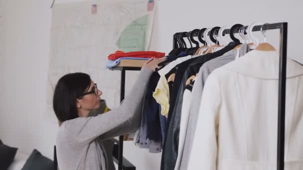 Designer de moda feminina trabalha em nova coleção para clientes em estúdio oficina aconchegante, costureira, alfaiate ou needlewoman de pé perto do rack de roupas com roupas elegantes na moda. Jovem morena — Vídeo de Stock