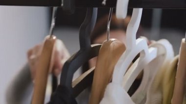 Yakın çekim bir kadeh bardaklarda renkli giysiler askıları, dokunmaktan ve onları hareket üzerinde düşünen genç kadın. Bayan giyim ön planda