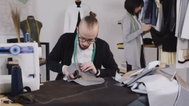 Sakallı studio işyerinde terzi ya da moda tasarımcısı oturur ve kesme gri kumaş büyük makas kullanarak. Arka planda, terzi elbiseler üzerinde hangi askıları görünüyor