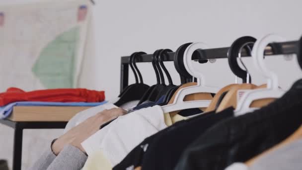 Close-up van een vrouwelijke handen lopen op kleding hangers spoor rek en op zoek naar de jurk op kleding winkel. Kaukasische jonge vrouw kijken naar mode kleding die opknoping op hangers. Kleerhangers — Stockvideo