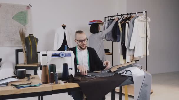 Sarto o stilista siede sul posto di lavoro in studio e il taglio di tessuto grigio con grandi forbici come segue i segni di gesso del modello utilizzando il modello. Sullo sfondo, la ragazza guarda — Video Stock