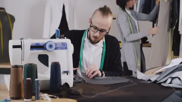 Handsome tailor sits on the workplace at studio and cutting gray fabric using large scissors as he follows the chalk markings of the pattern using pattern. In the background, the dressmaker looks at — Stock Video