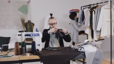 Terzi ya da moda tasarımcısı studio işyerinde oturur ve kesme gri kumaş büyük makas kullanarak. Arka planda, terzi beyaz elbise üzerinde hangi askı görünüyor