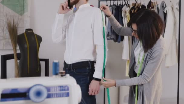 Young professional dressmaker taking measurements for sewing suit at tailors shop. Seamstress with measuring tape makes measurements for sewinf a new jacket for her client. Client with beard and — Stock Video