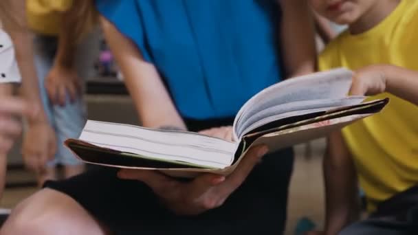 Καυκάσιος νηπιαγωγός και τους μαθητές της να καθίσει στο μαξιλάρια στο πάτωμα ο ίδιος, διαβάζοντας ένα βιβλίο μαζί σε μια τάξη. Δάσκαλος ανάγνωση του βιβλίου για μαθητές εξηγώντας το σχολικό μάθημα, να βοηθήσει με την εργασία — Αρχείο Βίντεο