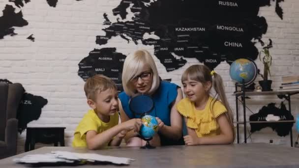 Profesor con dos niños en clase de geografía mirando al mundo. Lindo niño, niña y maestro mirando el mundo usando lupa en el aula en la escuela o jardín de infantes — Vídeo de stock