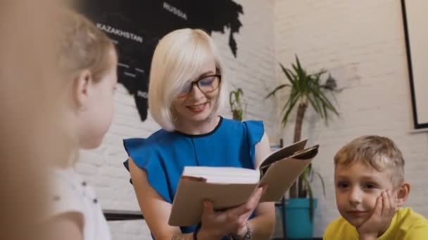 Mujer rubia joven en gafas de vista leer un libro para niños en la habitación en el fondo del mapa del mundo. Dos alumnos y un profesor leyendo el libro — Vídeos de Stock